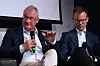 To menn sitter i en paneldebatt; den ene snakker i en mikrofon og gestikulerer.